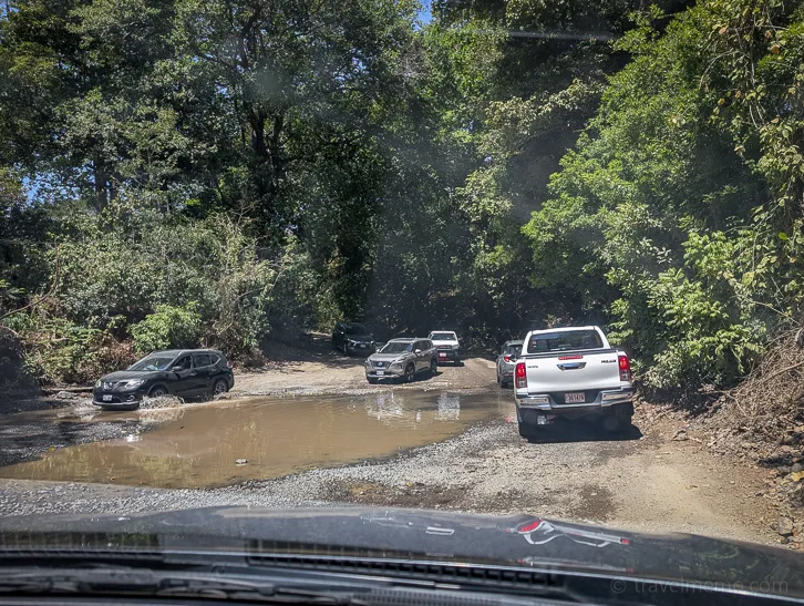 Several vehicles drive through a ford on a dirt road surrounded by dense green rainforest.
