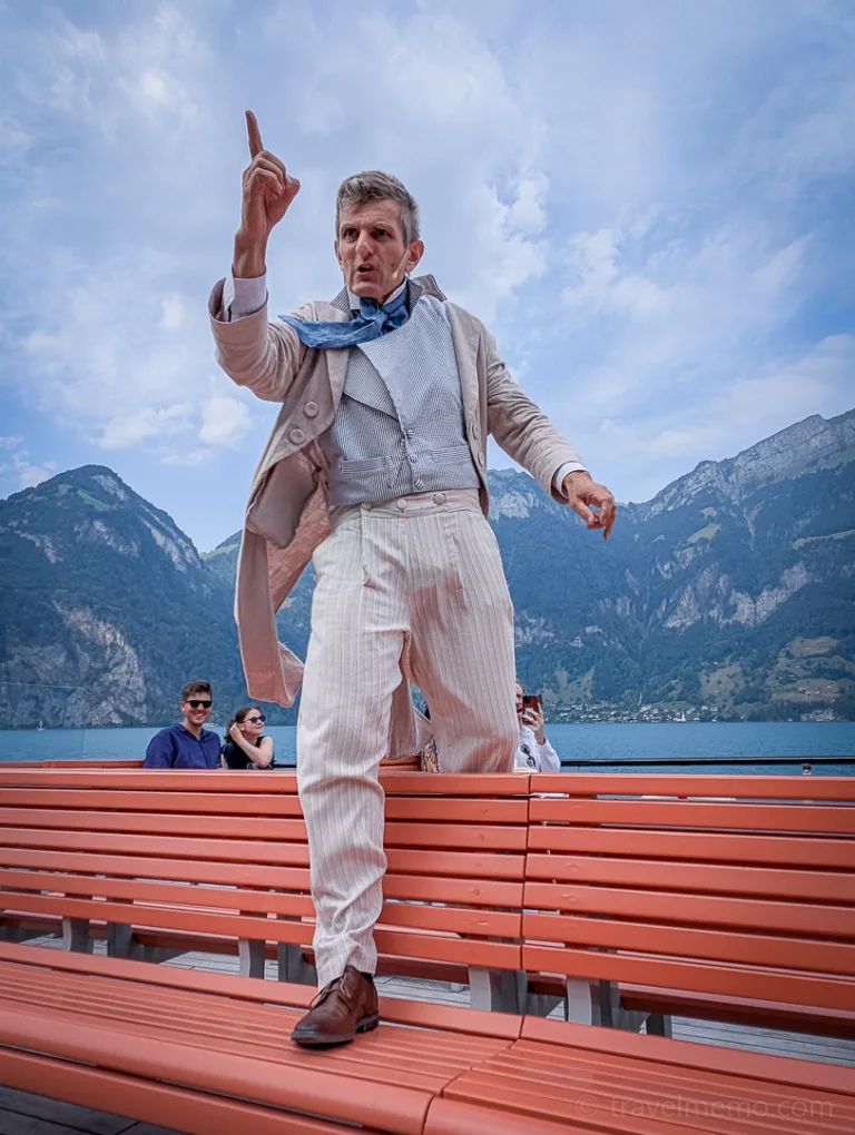 An actor dressed in light-colored clothing performs, gesturing, on orange benches in front of a lake and mountains