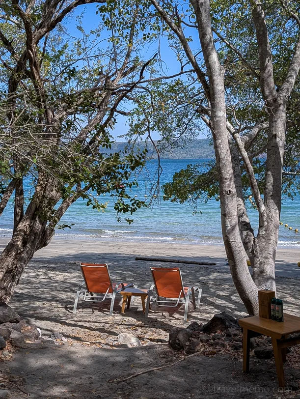 Two orange lounge chairs stand under large tropical trees right on the white sandy beach, with the turquoise-blue sea in the background