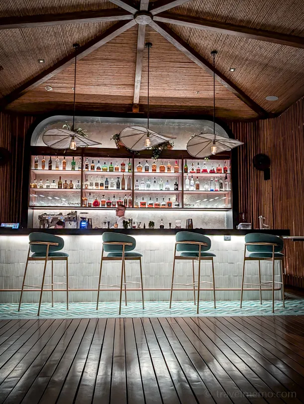 An elegant hotel bar with a wooden ceiling, four green bar stools, and an illuminated display of spirits in the background