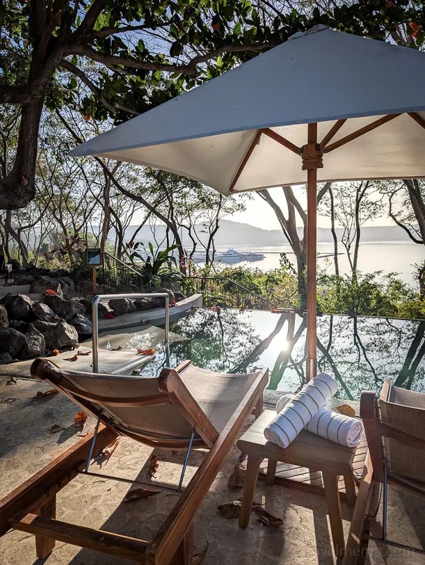Pool deck with lounge chairs and a parasol, surrounded by tropical vegetation and overlooking the sea