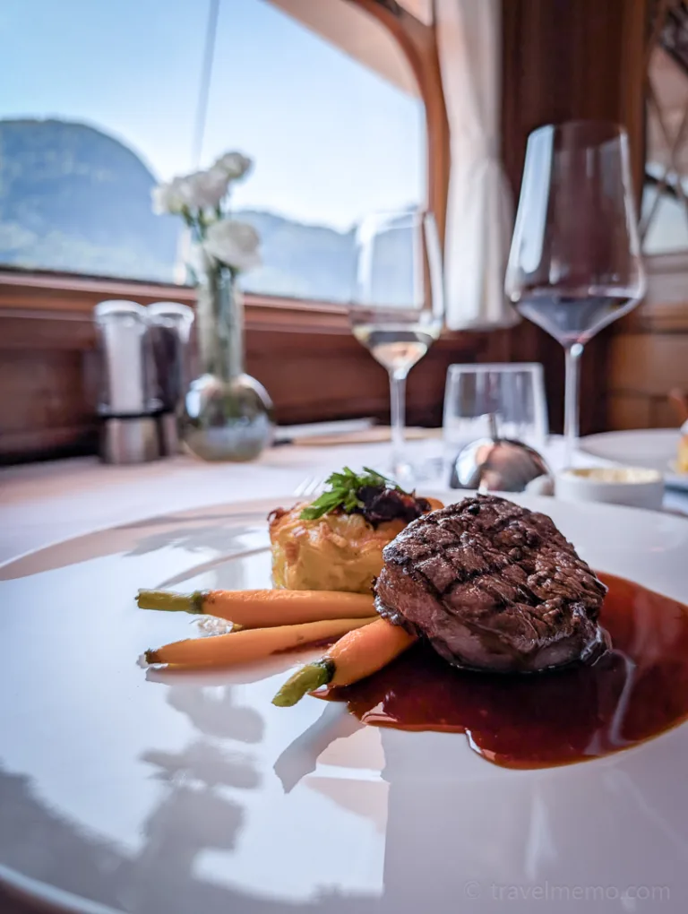 An elegant gourmet dish featuring vegetables and meat on a white plate, with a window in the background offering a view of the mountains