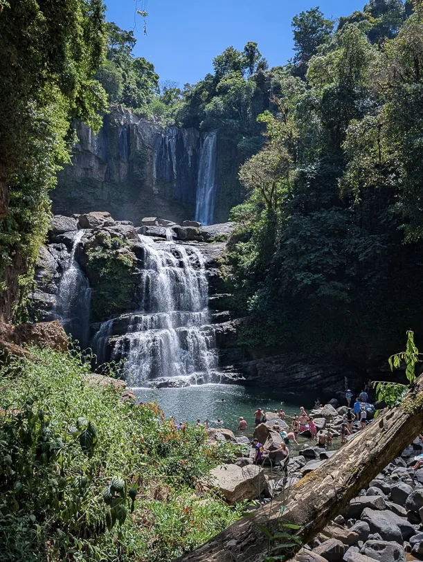A multi-tiered waterfall with a natural pool at its base, surrounded by dense tropical rainforest and visitors perched on the rocks