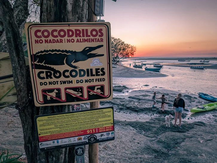 Warning sign with crocodile symbol and prohibition signs on Tamarindo beach at sunset, people and boats in the background
