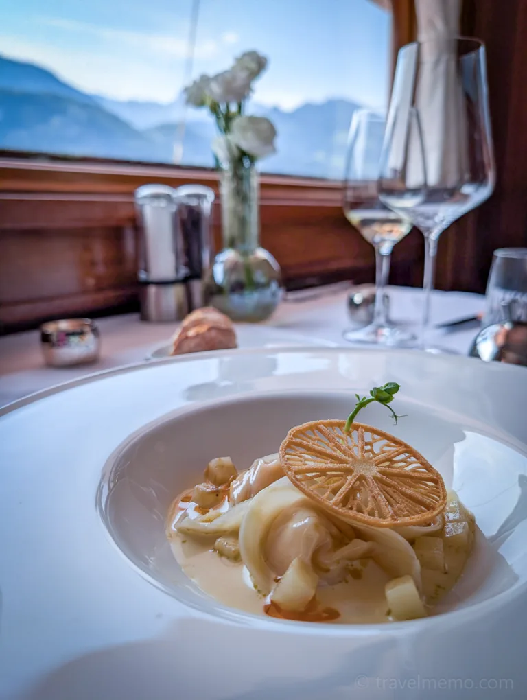 A table set in white with a gourmet main course served on a round plate, with a panoramic view of the mountains along Lake Lucerne in the background