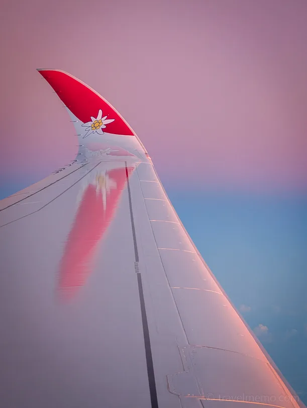 Wing of a Swiss Edelweiss airplane with red winglet and Edelweiss logo in front of a pink-purple evening sky.