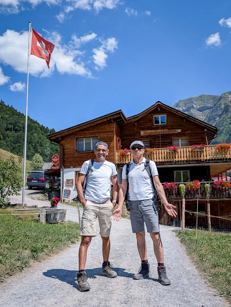 Two hikers are standing on a gravel path in front of a wooden chalet with a Swiss flag in an alpine landscape