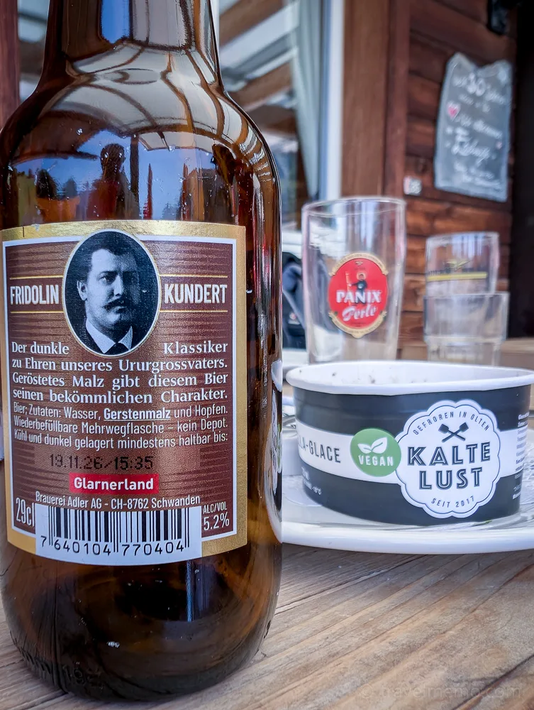 A beer bottle with a Fridolin Kundert label and a cup of vegan ice cream on a wooden table at a mountain restaurant