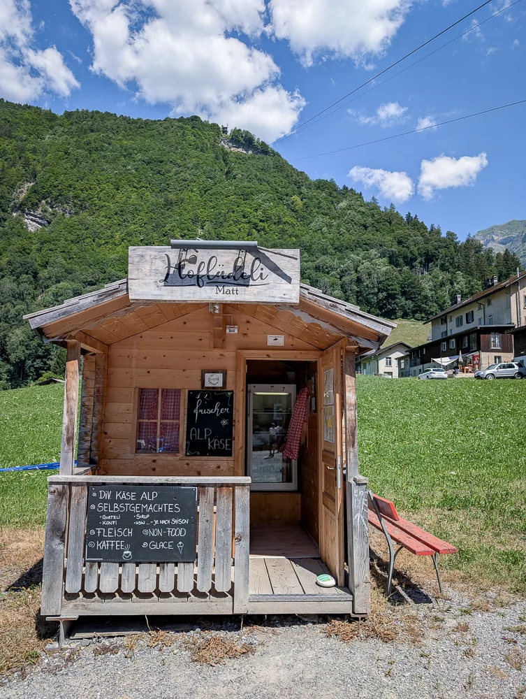 A small wooden farm stand with a display shelf set against a green mountain backdrop in Glarus South