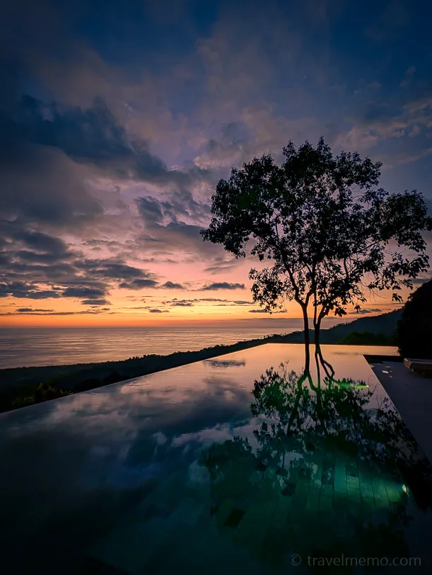 Kura Boutique Hotel infinity pool with a perfect reflection of a tree, the Pacific Ocean behind it and a dramatic sunset sky in orange and blue tones