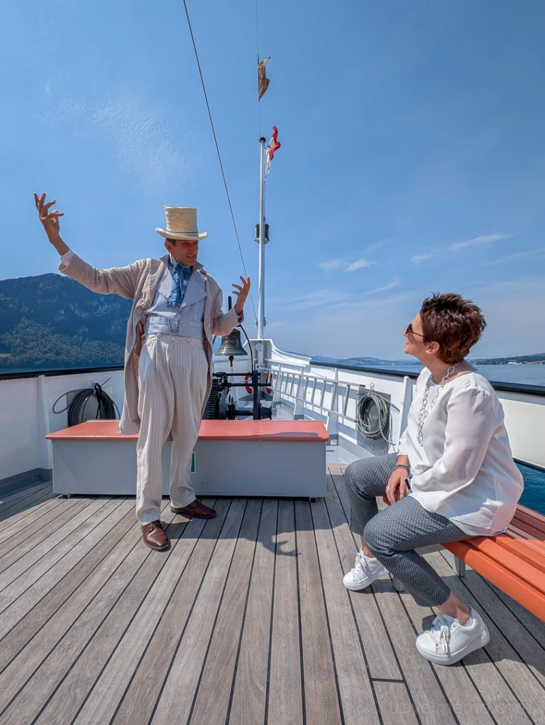An actor in period costume tells a story to a woman on the open deck of a steamboat on Lake Lucerne
