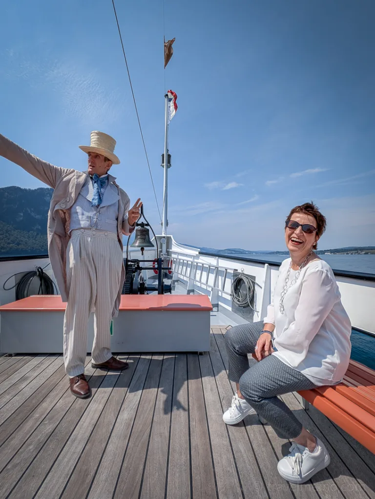 A woman laughs on a bench on the ship; next to her, a man in period clothing points out on the deck of the steamship