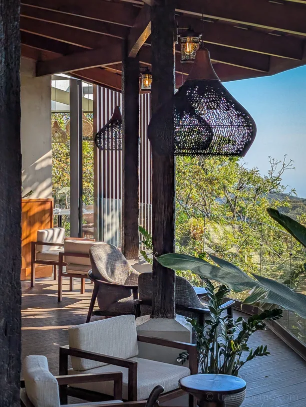Covered Koora hotel terrace with wooden furniture and large bamboo lamps in front of a tropical rainforest