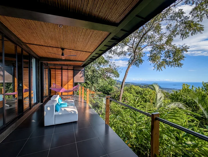 Covered hotel terrace of the Kura Boutique Hotel with white furniture and panoramic views over tropical rainforest to the Pacific Ocean.