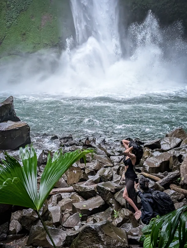 A hiker stands on boulders in front of a high waterfall, surrounded by tropical vegetation and dense spray mist