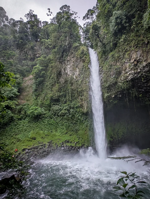 A high waterfall plunges through dense tropical rainforest into a turquoise pool surrounded by lush green vegetation