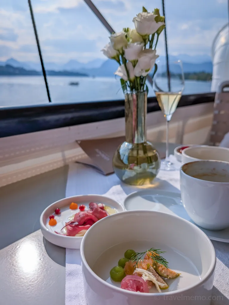 An elegantly plated gourmet dish and coffee on a white table with a view of the lake, with flowers in a vase and a champagne glass in the background