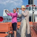 Two men on the deck of a steamboat, one in historical costume; both are looking through telescopes