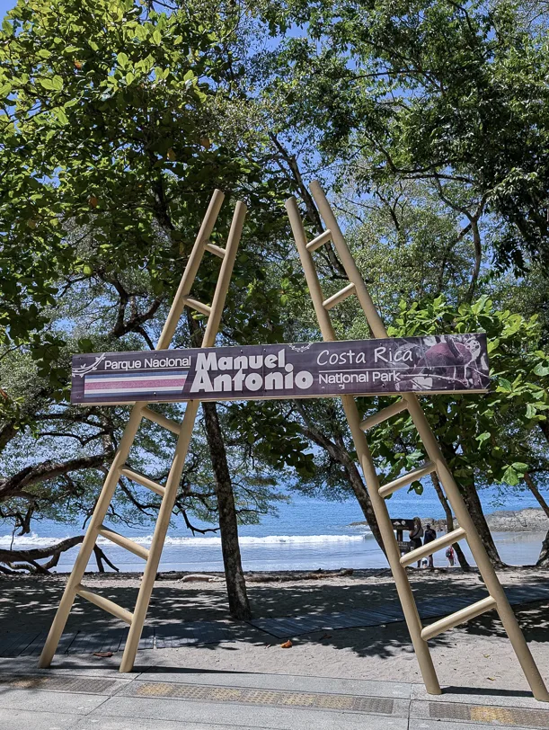 Wooden sign of the Parque Nacional Manuel Antonio under green trees with a view of the white sandy beach and blue sea