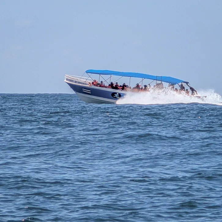 A white speedboat with a blue sunroof travels at high speed across the deep blue sea of the Marino Ballena National Park, creating a large cloud of spray.