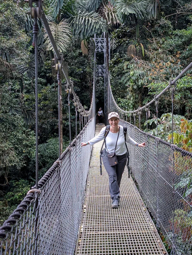 Hiker crosses a metal suspension bridge between tall trees in the tropical rainforest
