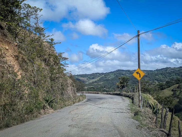 Unpaved gravel road with curve warning leads through green mountain landscape in Costa Rica