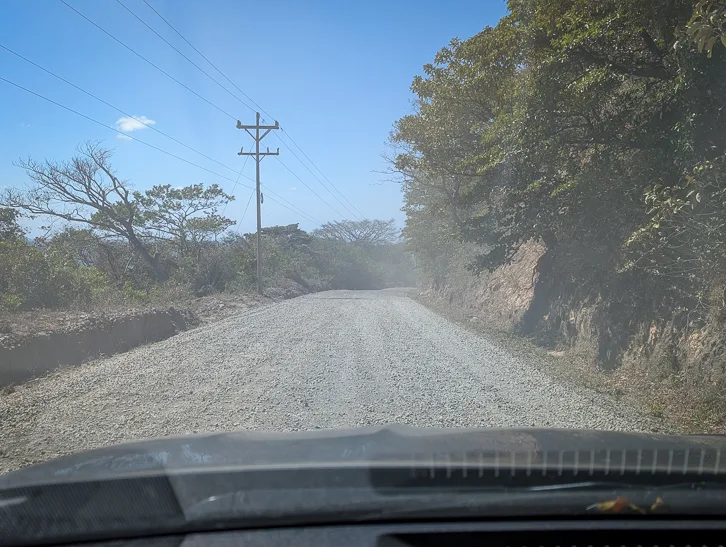 View through the windshield of a dusty gravel road with power lines and tropical vegetation on the sides