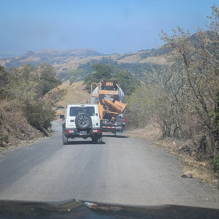 View of a mountain road with a white off-road vehicle and a truck, surrounded by wooded hills under a blue sky