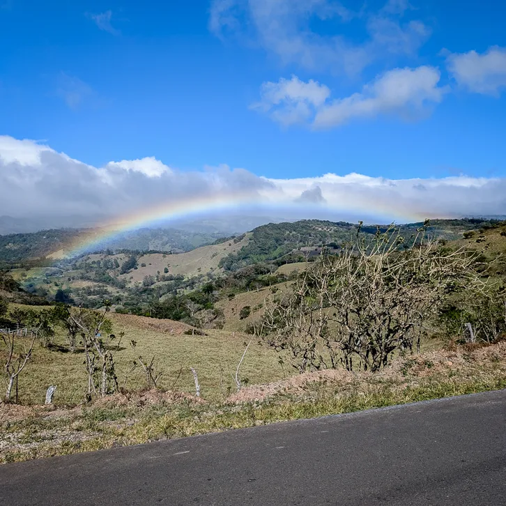 A brilliant rainbow stretches across the green hills of Monteverde under a partly cloudy blue sky