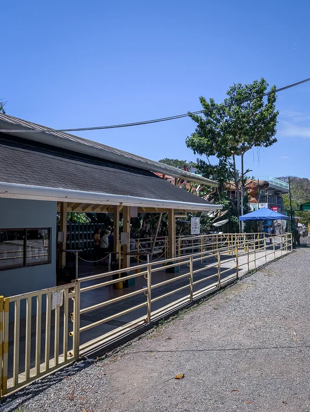 Entrance area of Manuel Antonio National Park with roofed building, wooden railings and tropical vegetation under a blue sky