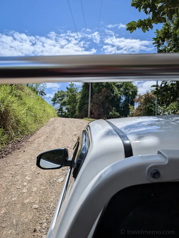 A silver car on an unpaved gravel road, surrounded by dense green tropical forest under a blue sky dotted with white clouds
