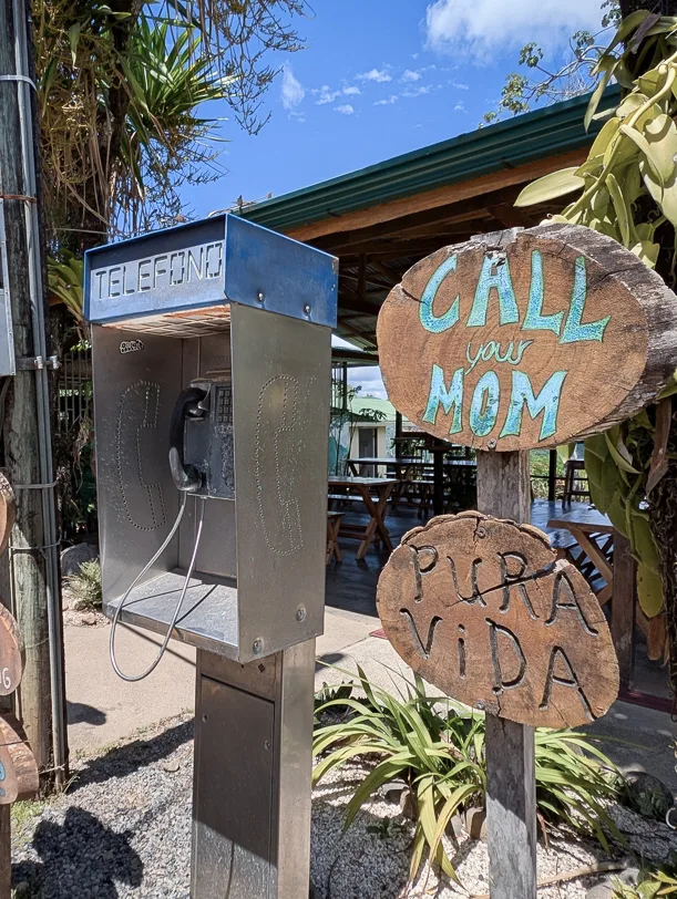 An old phone booth with wooden signs reading 'Call your Mom' and 'Pura Vida' set against a backdrop of tropical vegetation