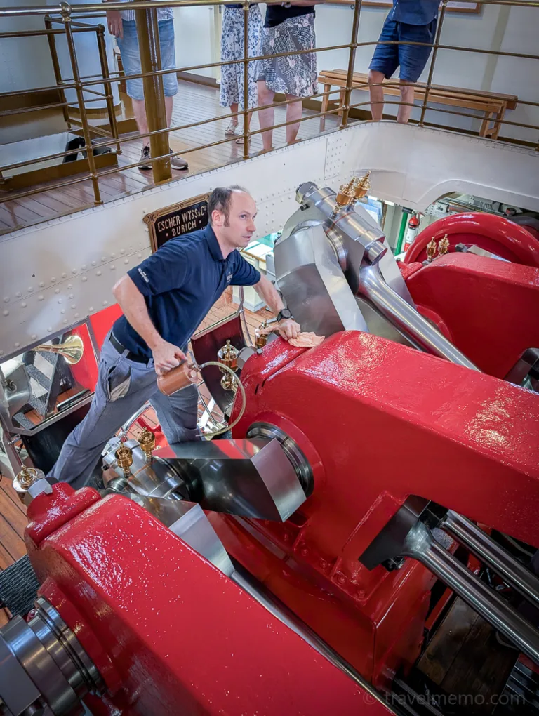 A man operates the large red engines of a historic steamship in the engine room, surrounded by spectators on the gallery