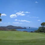 Gepflegtes Golfgrün mit Flagge im Vordergrund, dahinter blaues Meer und bewaldete Hügel unter blauem Himmel mit weissen Wolken