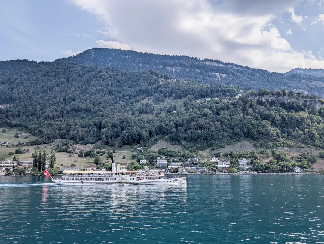 A steamboat flying the Swiss flag sails on Lake Lucerne off the town of Weggis, with green mountain slopes in the background