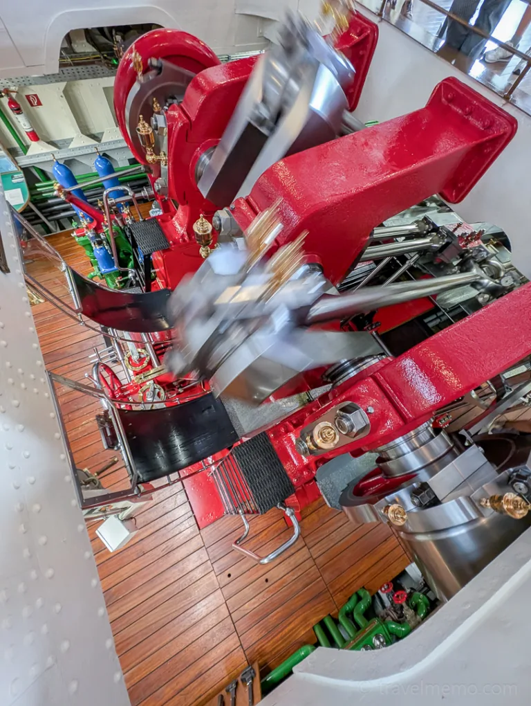 A view into the running engine room of a red-painted paddle steamer with a wooden deck