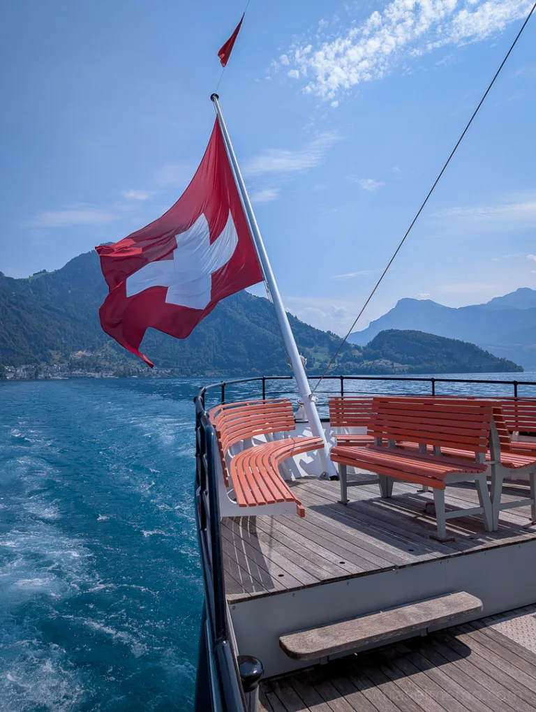 The stern of a sightseeing boat flying the Swiss flag, with empty benches, on Lake Lucerne, surrounded by mountains
