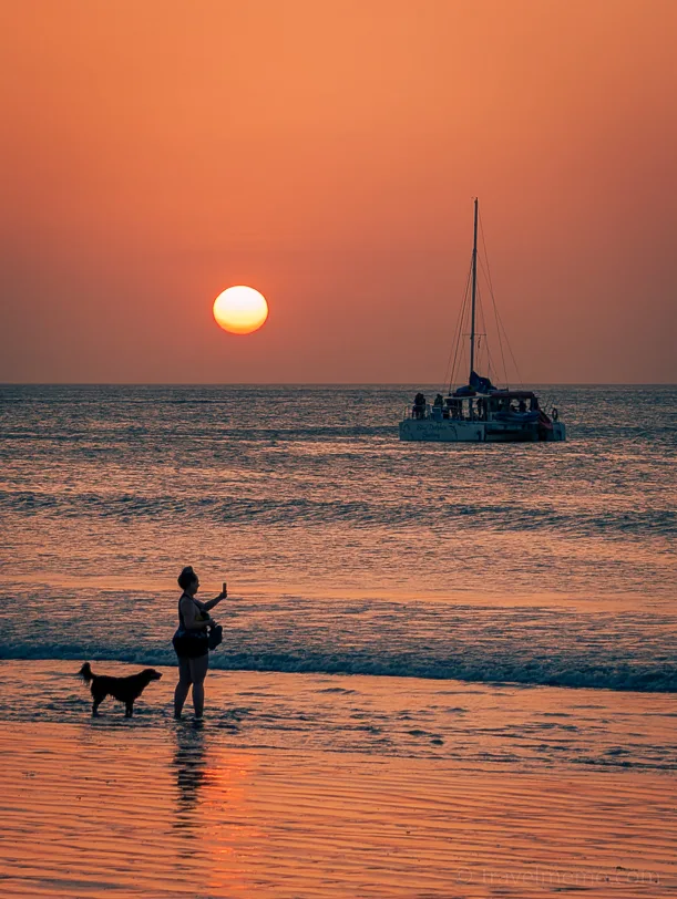 Silhouette of a person with a dog on the beach at sunset, in the background a sailing boat on the sea.