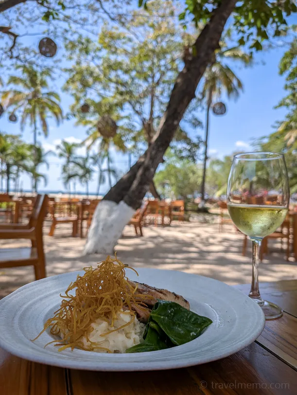 White plate with mashed potatoes, fish and green vegetables on a wooden table, a glass of white wine next to it, tropical palm trees and a beach restaurant atmosphere in the background