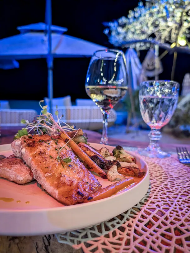 Grilled fish on a white plate with white wine and a glass of water on the woven table surface of an illuminated restaurant terrace