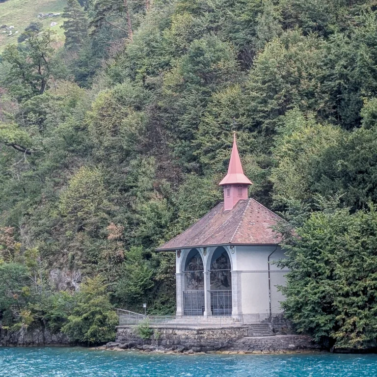 A small chapel with a pointed tower stands on the shore of the turquoise-blue Lake Lucerne, set against a dense forest