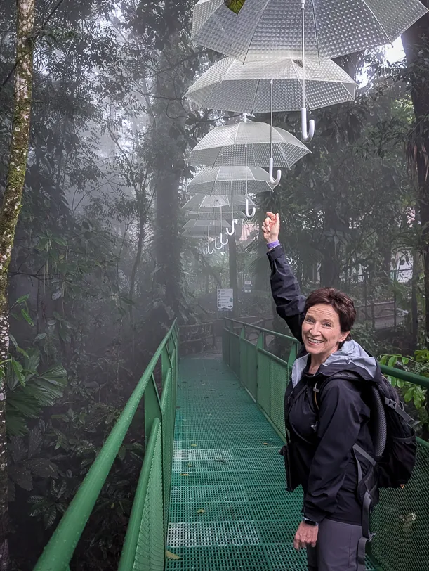 Hiker on a green suspension bridge under an artistic installation of white umbrellas in a misty tropical rainforest