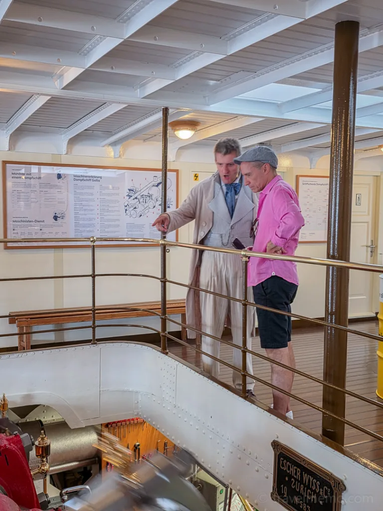 Two men are standing on the deck of a historic steamboat, looking into the engine room