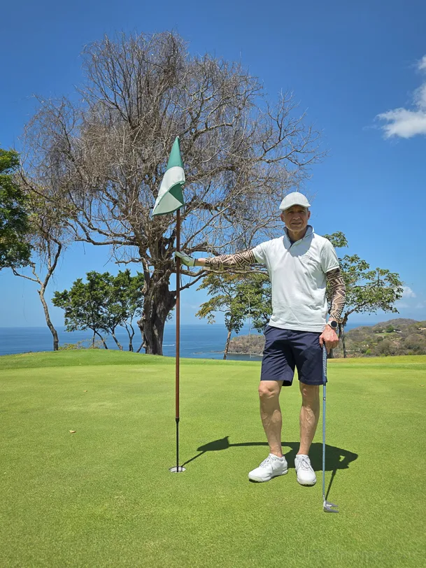 A golfer in white stands with a club next to a flag on a green golf course with a view of the sea and a blue sky
