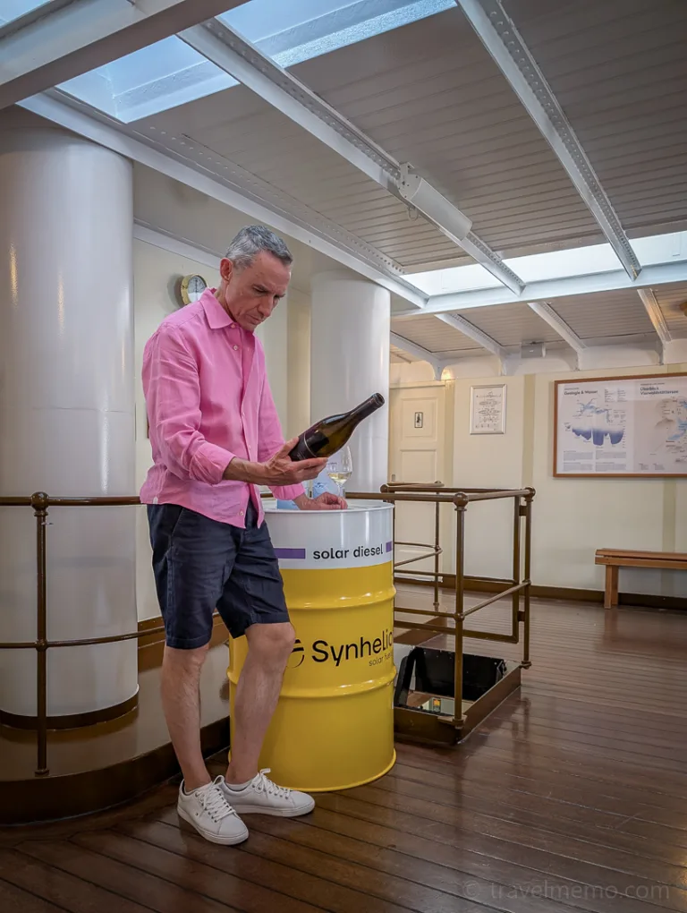 A man in a pink shirt looks at a bottle next to a yellow barrel inside a historic steamboat on Lake Lucerne